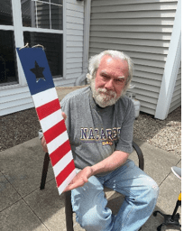 An older man with white hair and a beard sits outside, holding a wooden plank painted with red and white stripes and a blue section with a black star. He wears a gray NIAGARA T-shirt and jeans.