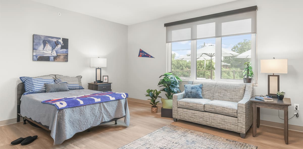 Bed with Buffalo Bills blanket, couch, and table with lamp with windows in the background