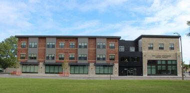 A three-story brick and stone building, labeled True North Apartments Dental Avenue, with large windows on the ground floor and a green lawn in front.