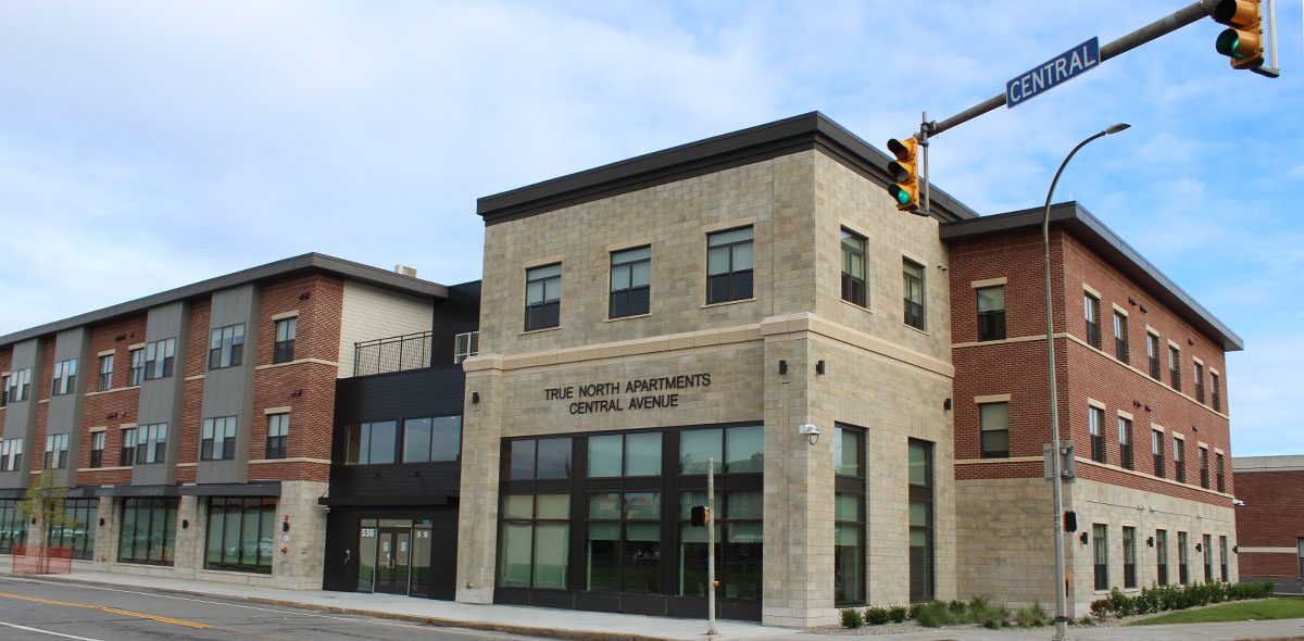A modern three-story apartment building with brick and stone exterior, large windows, and a sign reading True North Apartments Central Avenue at a street corner with a traffic light and street sign reading Central..