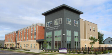 A modern three-story apartment building with a mix of gray, beige, and red brick exterior. A sign reads True North Apartments Portland Avenue. Trees and a black fence line the sidewalk in front.