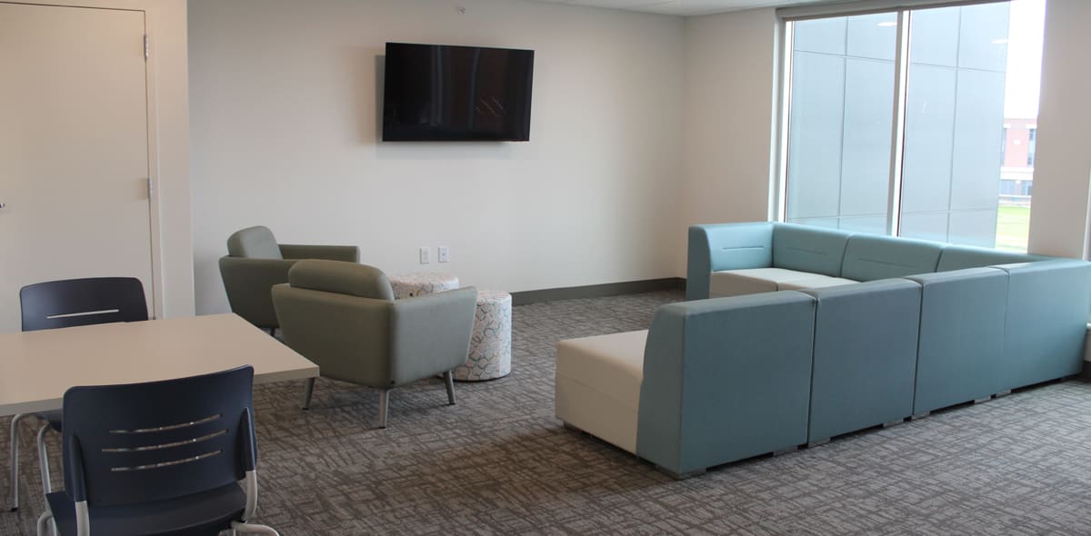 A modern lounge with a blue sectional sofa, two armchairs, a wall-mounted TV, and a table with chairs. Large windows let in natural light.