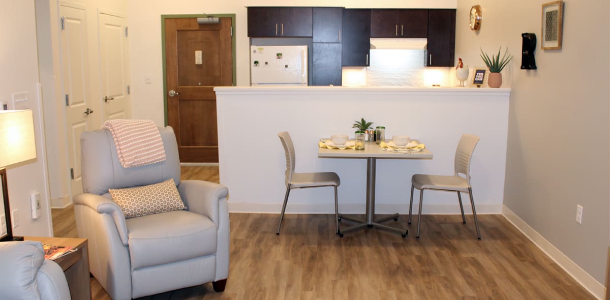 A cozy apartment living area with a recliner, small dining table set for two, and an open kitchen with dark cabinets and a white fridge, all on wood flooring.