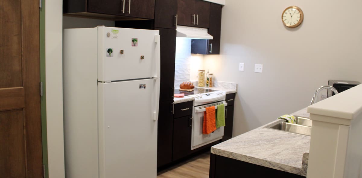 Small kitchen with dark cabinets, white refrigerator and stove, orange and green towels on oven, marble counter, sink, and a wall clock. Light wood floor and closed wooden door are also visible.