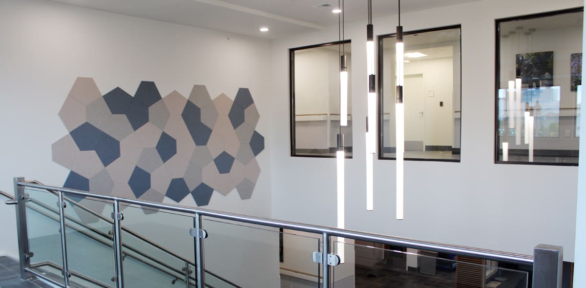 Modern interior with a glass staircase railing, geometric wall art in neutral colors, hanging tube lights, and windows looking into a bright office space.