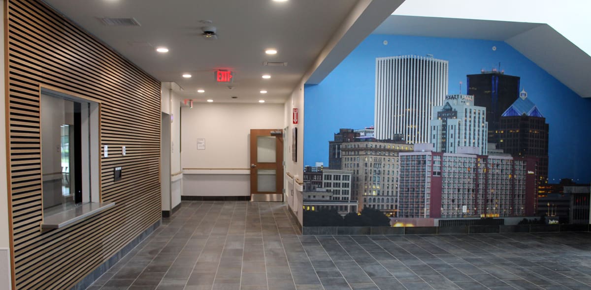 A hallway with dark tile flooring, wooden wall panels, a reception window, and a mural of a city skyline on the right wall under a slanted ceiling.