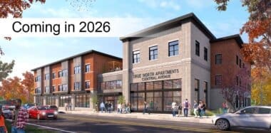 A modern three-story apartment building with a sign reading True North Apartments Central Avenue. People walk on the sidewalk, and cars are parked along the street. Text overlay says Coming in 2026. Trees with autumn leaves surround the scene.