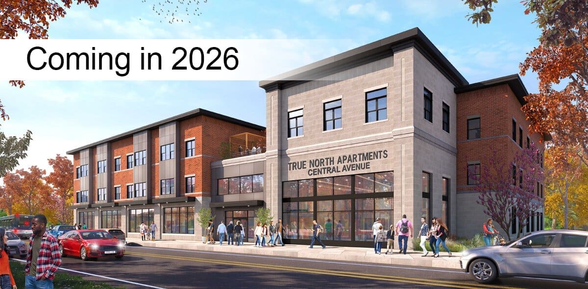 A modern three-story apartment building with a sign reading True North Apartments Central Avenue. People walk on the sidewalk, and cars are parked along the street. Text overlay says Coming in 2026. Trees with autumn leaves surround the scene.