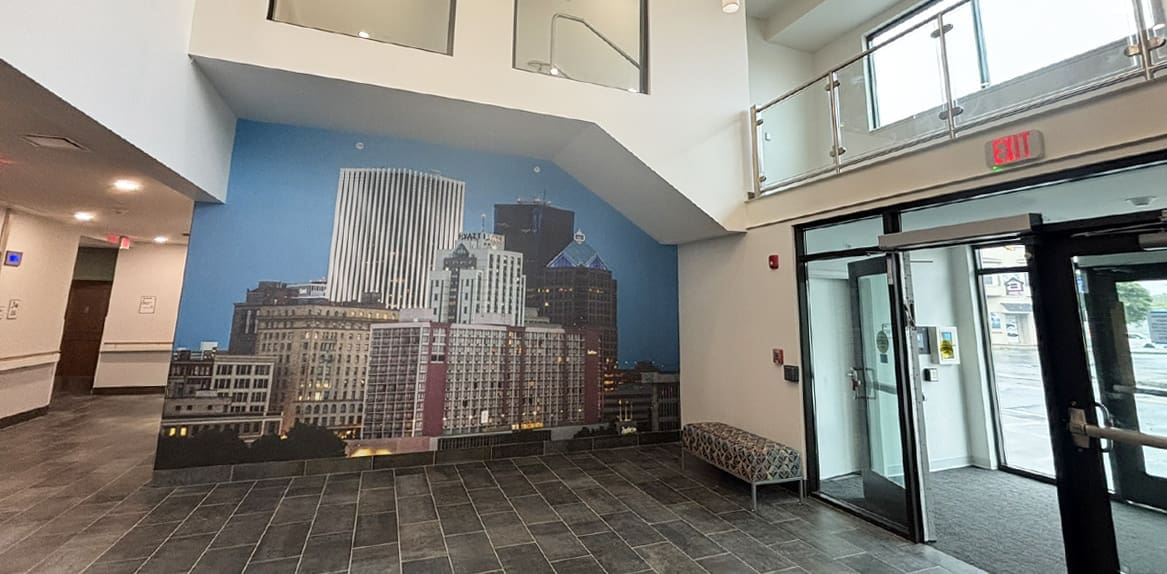 A modern lobby with gray tile floors, glass doors, a patterned bench, and a large wall mural depicting city skyscrapers beneath a stair landing.