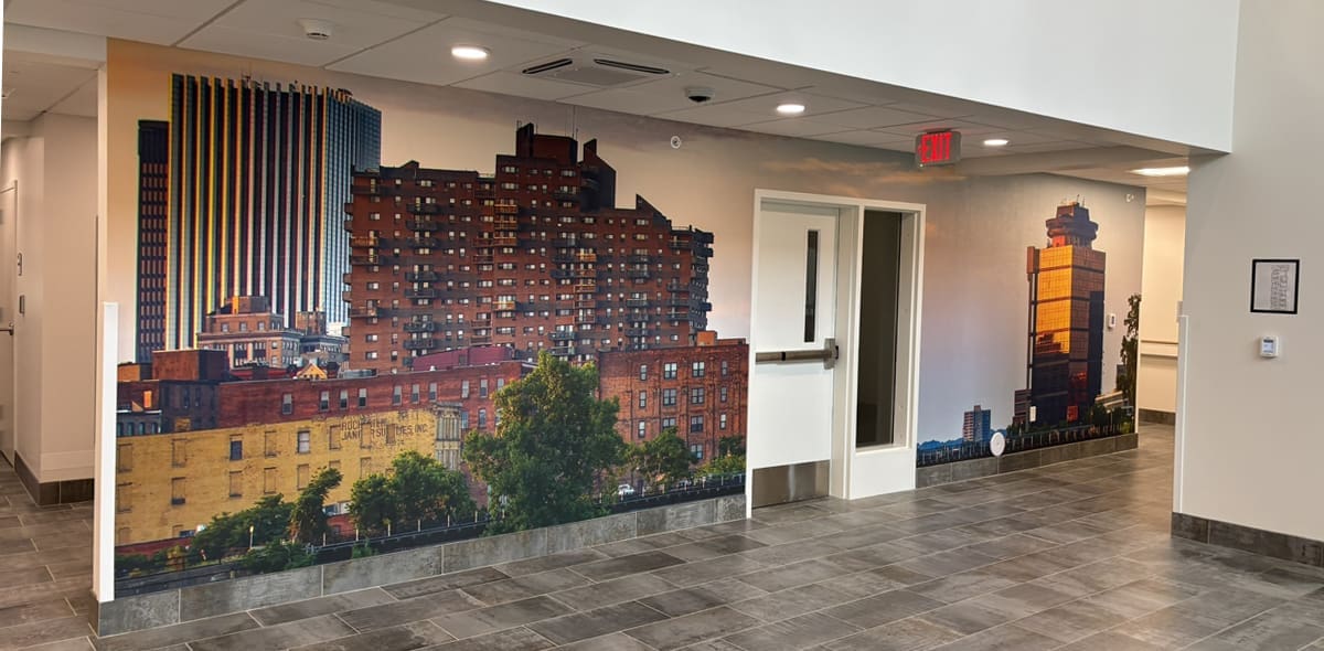 A wall mural in a modern building lobby depicts tall city buildings, trees, and a clear sky, creating an urban cityscape scene across the walls near a doorway and hallway.
