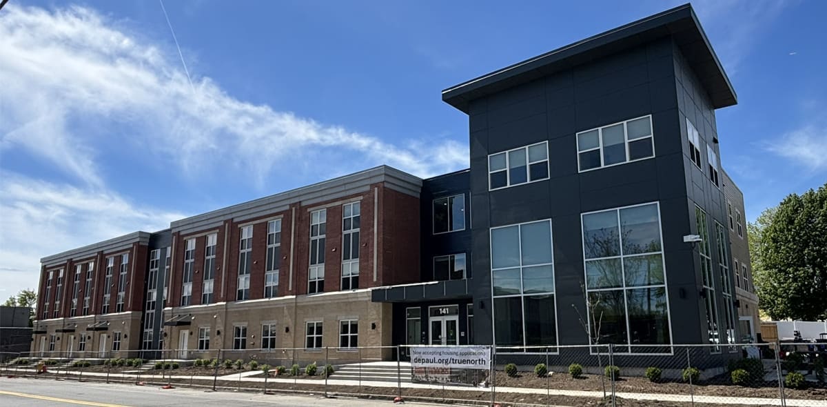 A modern three-story building with red brick and dark gray panels, large windows, and a fenced-off area in front. A banner hangs on the fence, and trees are visible in the background under a partly cloudy sky.