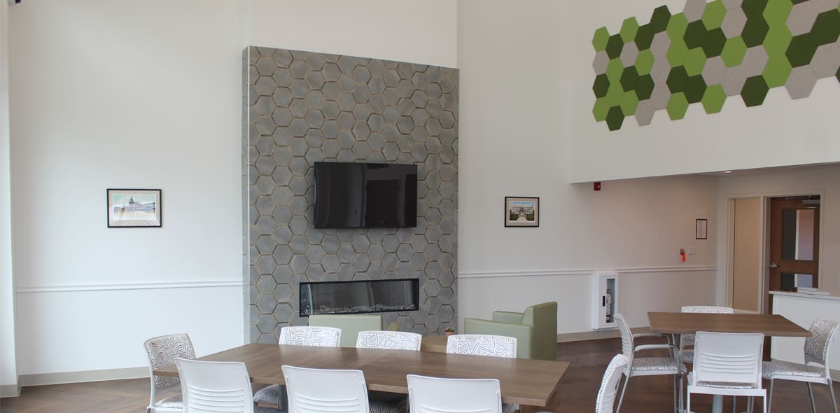 A modern lounge with wooden tables, white chairs, a TV mounted above a fireplace, and a green hexagonal wall art display. Large windows provide natural light.