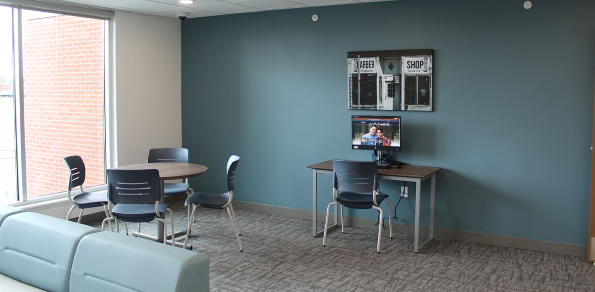 A modern lounge with a round table and four chairs, a desk with a computer, and large windows. The walls are blue and beige, and the floor is carpeted.