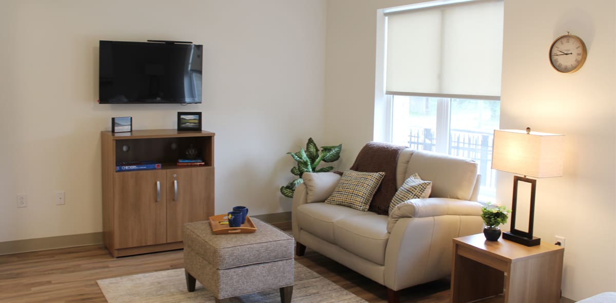 A cozy living room with a loveseat, ottoman, lamp, potted plant, and a TV mounted on the wall above a wooden cabinet. A clock hangs near a window with blinds letting in daylight.