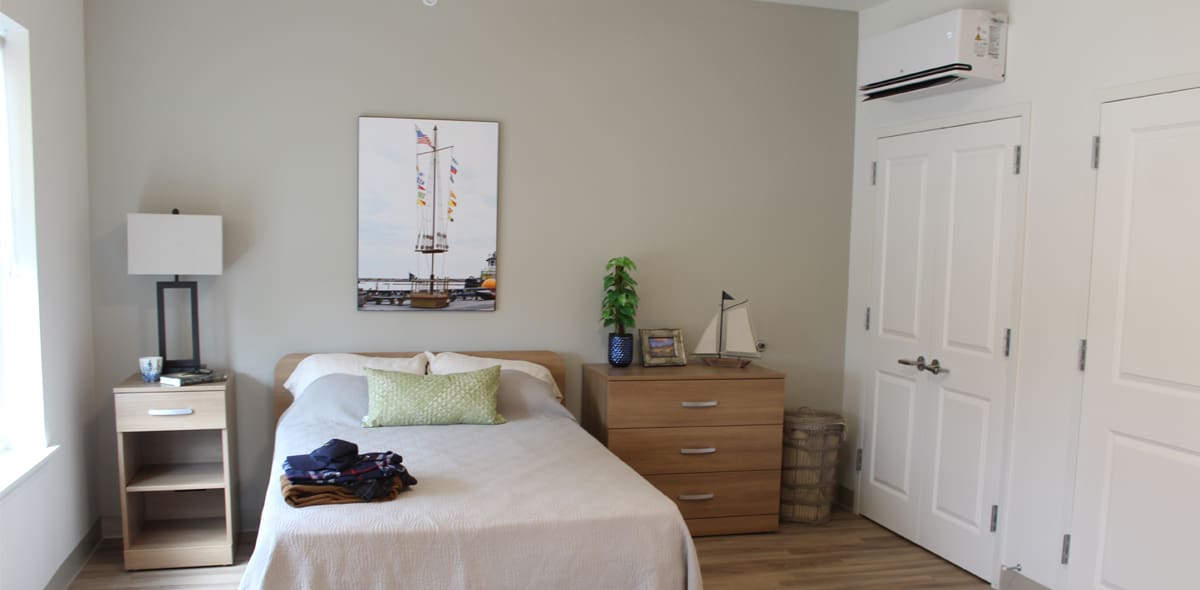 A neatly made bed with a green pillow is beside a wooden nightstand and dresser. A lamp, plant, photo, and sailboat decor are on the dresser. A sailboat picture hangs above the bed. White double doors are on the right.