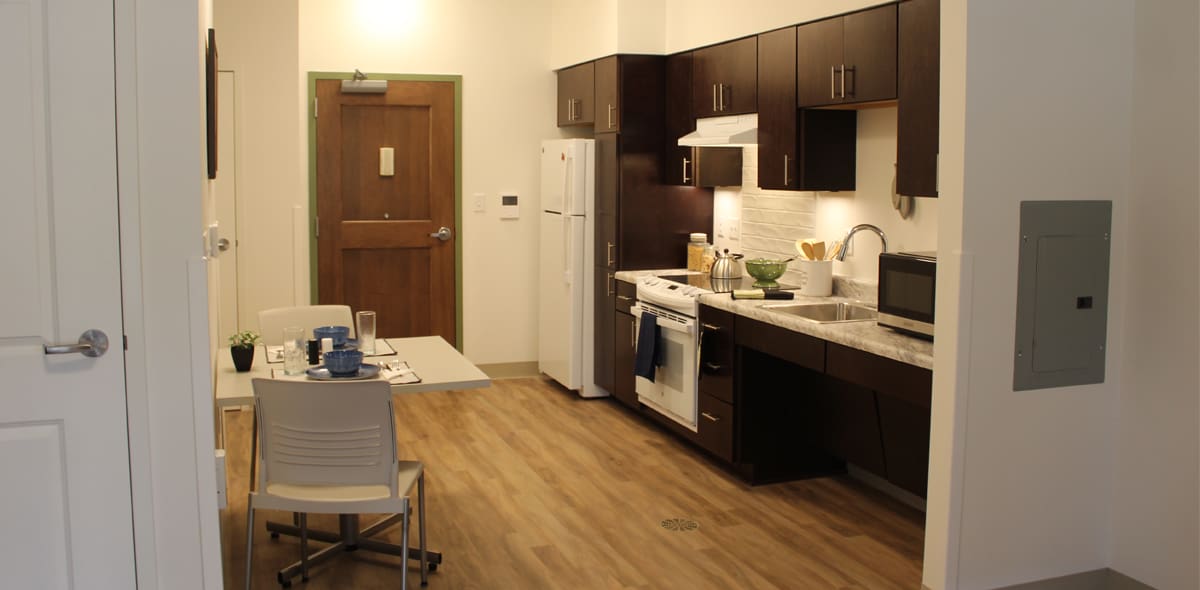 A modern, compact kitchen with dark cabinets, white appliances, and a small dining table set for two on wood flooring. A closed wooden door is visible at the back.