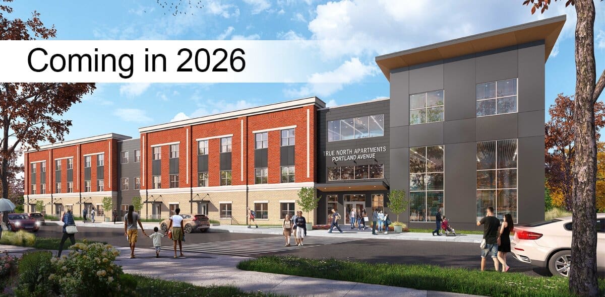 A modern three-story apartment building with red and gray exterior, people walking nearby, and a sign reading Coming in 2026 across the top left of the image.