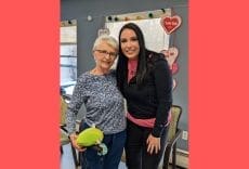 Two women smile and pose together indoors. One woman holds a green plush toy, and there are heart decorations on the wall behind them. The background has windows and chairs, and the image has a red border.