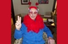 A person wearing heart-shaped glasses, red heart antennae, and a red feather boa makes a peace sign while sitting indoors, smiling at the camera.