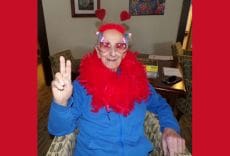 A person wearing heart-shaped glasses, red heart antennae, and a red feather boa makes a peace sign while sitting indoors, smiling at the camera.