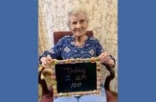 An elderly woman with short gray hair sits in a chair, smiling and holding a chalkboard that reads, Today I am 100. The background features a decorative cream-colored curtain.