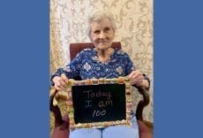 An elderly woman with short gray hair sits in a chair, smiling and holding a chalkboard that reads, Today I am 100. The background features a decorative cream-colored curtain.