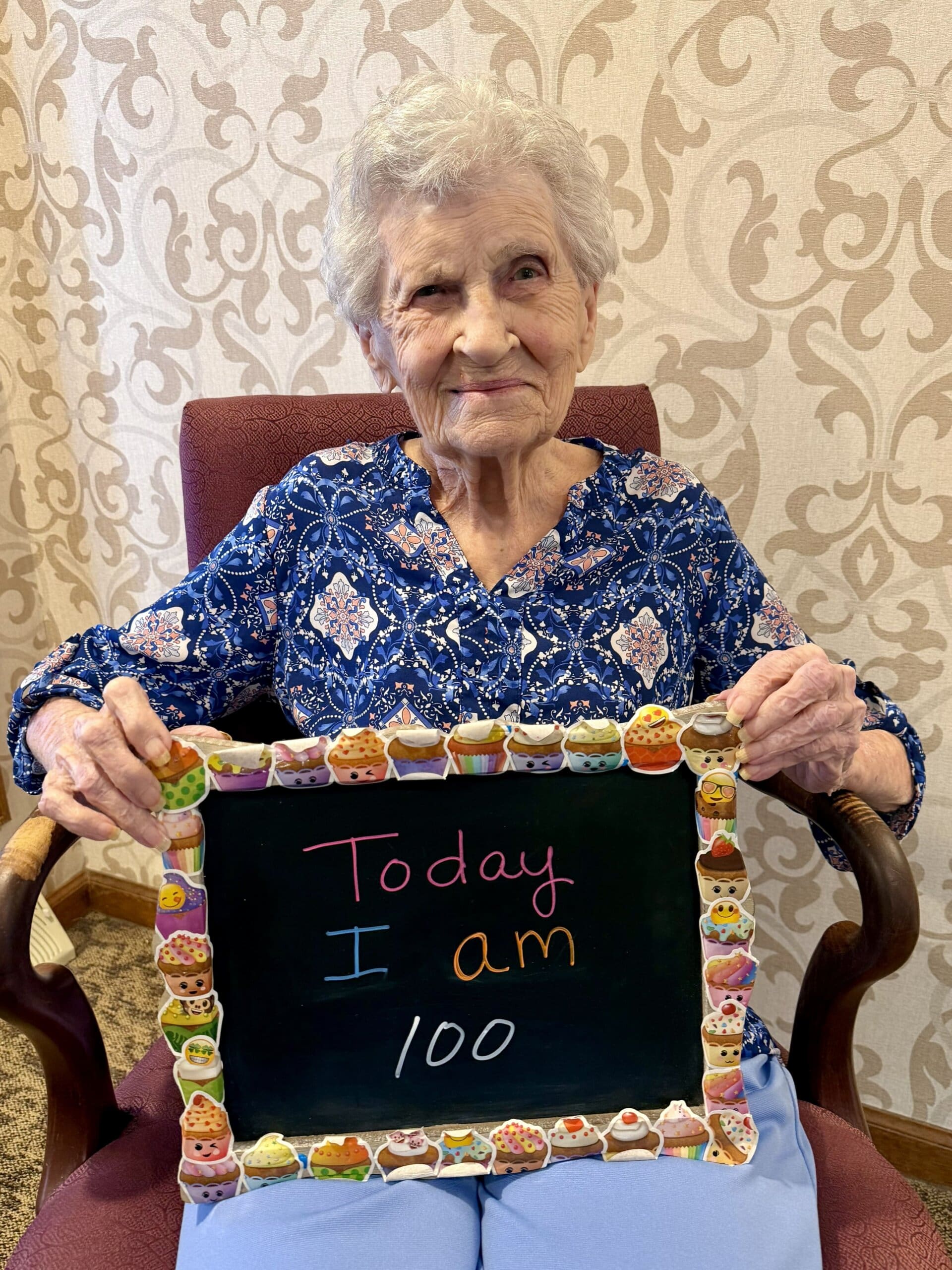 An elderly woman with short gray hair sits on a chair, smiling and holding a decorated chalkboard that reads Today I am 100 in colorful letters. The background features a beige patterned wall.