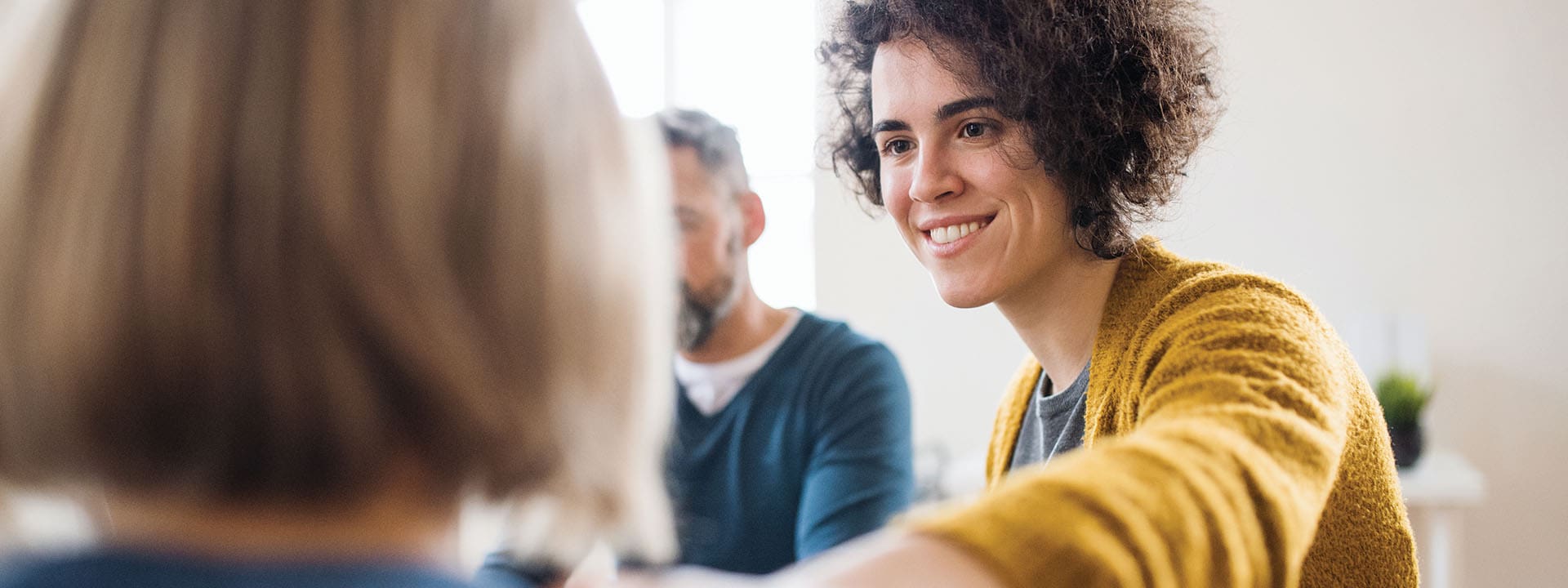 A woman with curly hair and a yellow sweater smiles and reaches out to another person in the foreground during a friendly conversation, with a man sitting in the background.