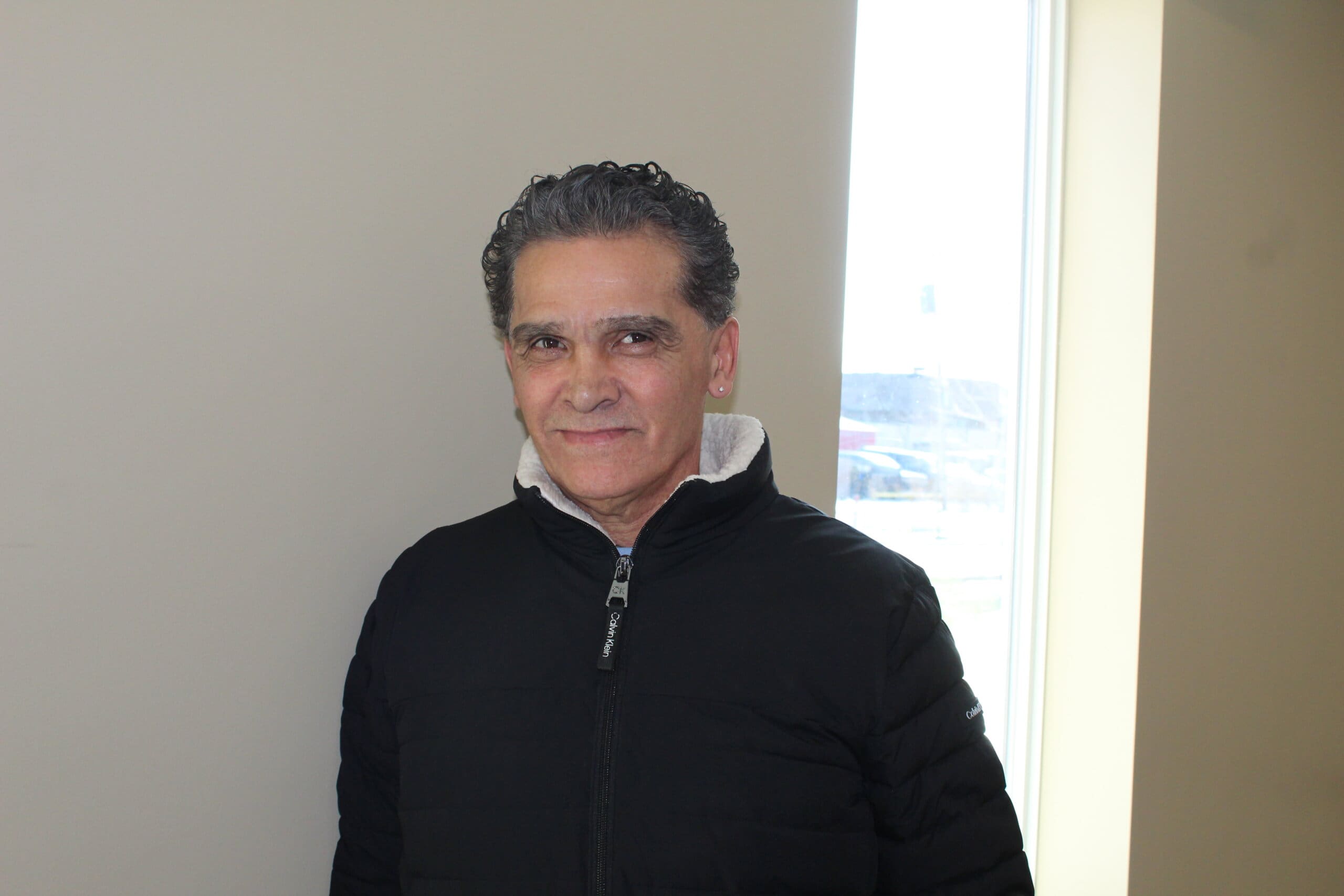 A person with short dark hair and a black zip-up jacket stands indoors near a window, smiling slightly. Natural light comes in from the window on his right side.