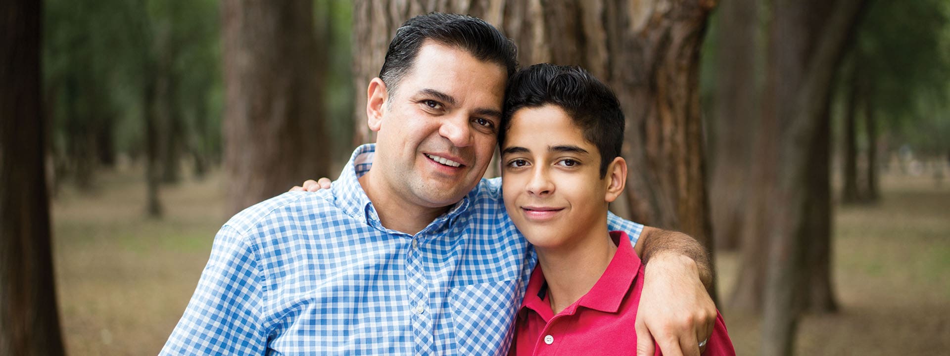 A smiling man in a blue checkered shirt hugs a boy in a red polo shirt, both standing closely together in a wooded outdoor setting.