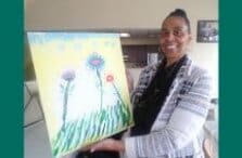 A smiling woman holds up a colorful painting of flowers with green stems, a yellow background, and a blue sky, standing indoors.