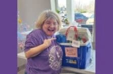 A smiling woman with blonde hair and glasses wearing a purple shirt points excitedly at a blue cooler with a sign that says Buddy Basket and tags attached, indoors near a window.