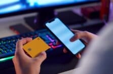 A person holds a yellow contactless credit card and a smartphone in front of a computer keyboard with colorful backlighting, suggesting an online payment or banking activity.