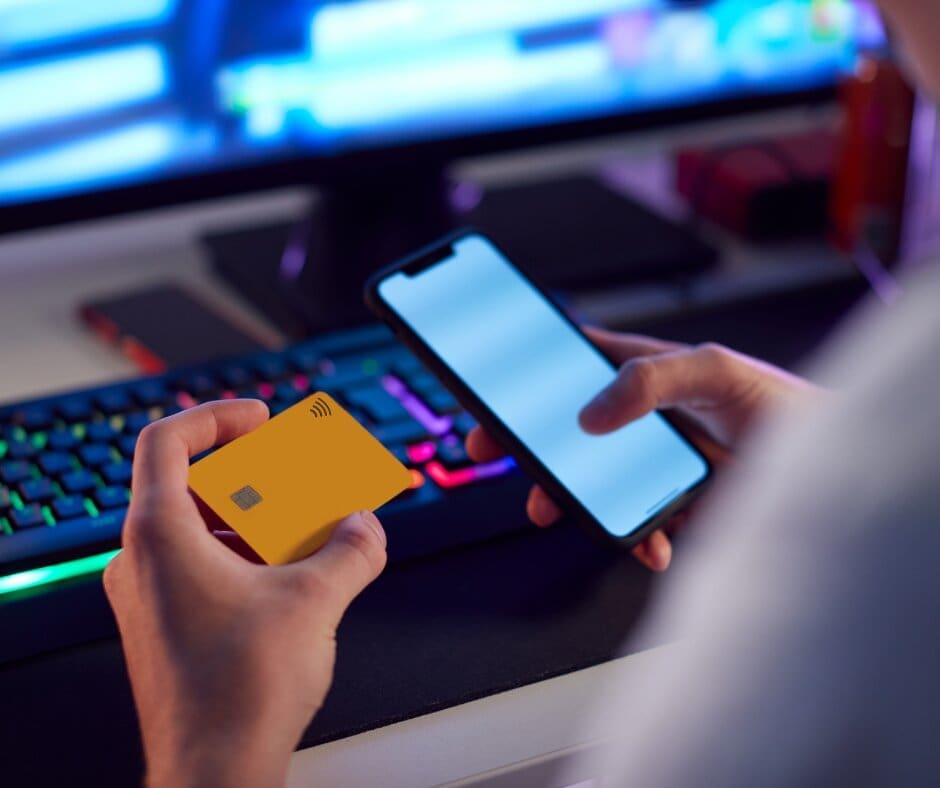 A person holds a yellow contactless credit card and a smartphone in front of a computer keyboard with colorful backlighting, suggesting an online payment or banking activity.