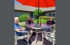 Three older adults sit around a patio table under a large red umbrella, enjoying a sunny day outside a building with a garden and flowers nearby.