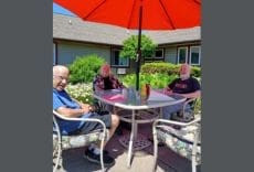 Three older adults sit around a patio table under a large red umbrella, enjoying a sunny day outside a building with a garden and flowers nearby.