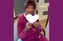 An elderly woman in a burgundy tracksuit sits on a chair, smiling and holding a white paper heart. The background features a wall with white trim, and purple borders frame the image.