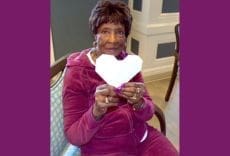 An elderly woman in a burgundy tracksuit sits on a chair, smiling and holding a white paper heart. The background features a wall with white trim, and purple borders frame the image.