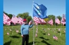 A person wearing a red hat and blue sweater stands outdoors on grass, holding a blue flag among many American flags displayed on poles under a clear sky.