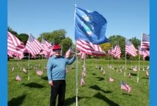 A person wearing a red hat and blue sweater stands outdoors on grass, holding a blue flag among many American flags displayed on poles under a clear sky.