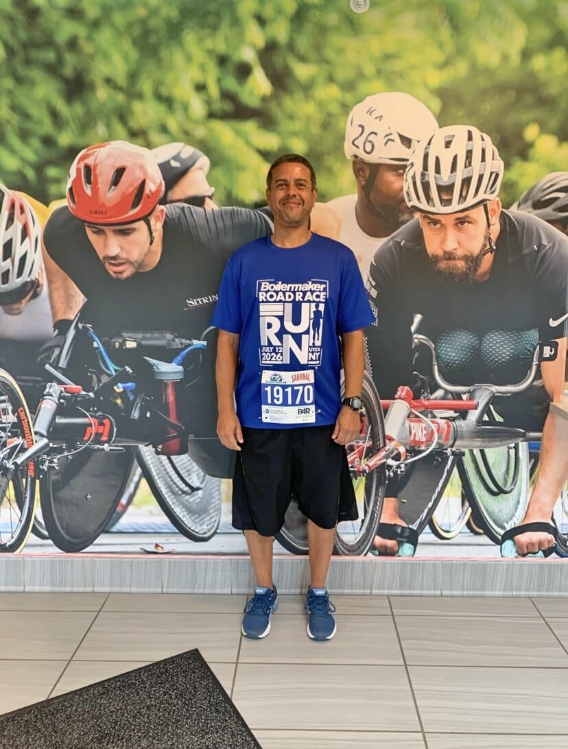 A man stands indoors, smiling, wearing a Boilermaker Road Race shirt and bib. Behind him is a wall mural showing athletes racing in wheelchairs, wearing helmets and athletic gear.