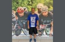 A man in sportswear stands indoors in front of a wall mural showing three cyclists racing, wearing helmets and focused expressions.