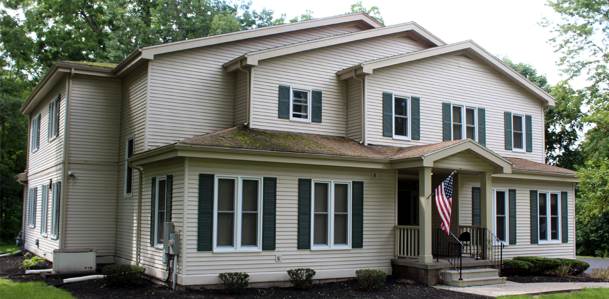 Two-story beige house with green shutters, multiple windows, and a covered front porch displaying an American flag. Surrounded by trees and small bushes, the house has a dark roof and a well-kept front yard.