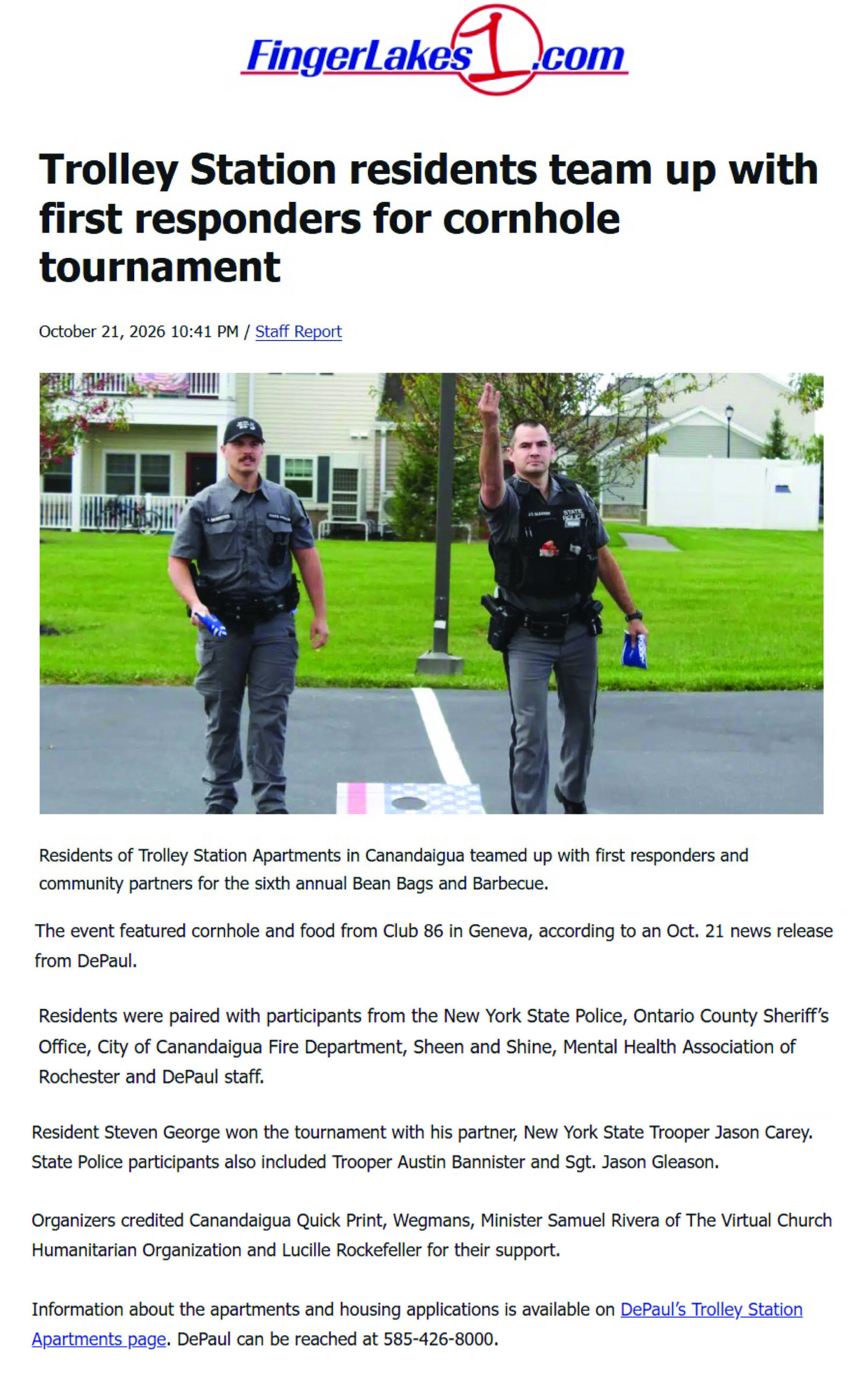 Two New York State Troopers standing on a lawn, one gesturing with his hand and holding a bean bag, in front of the FingerLakes1.com news article about a cornhole tournament.